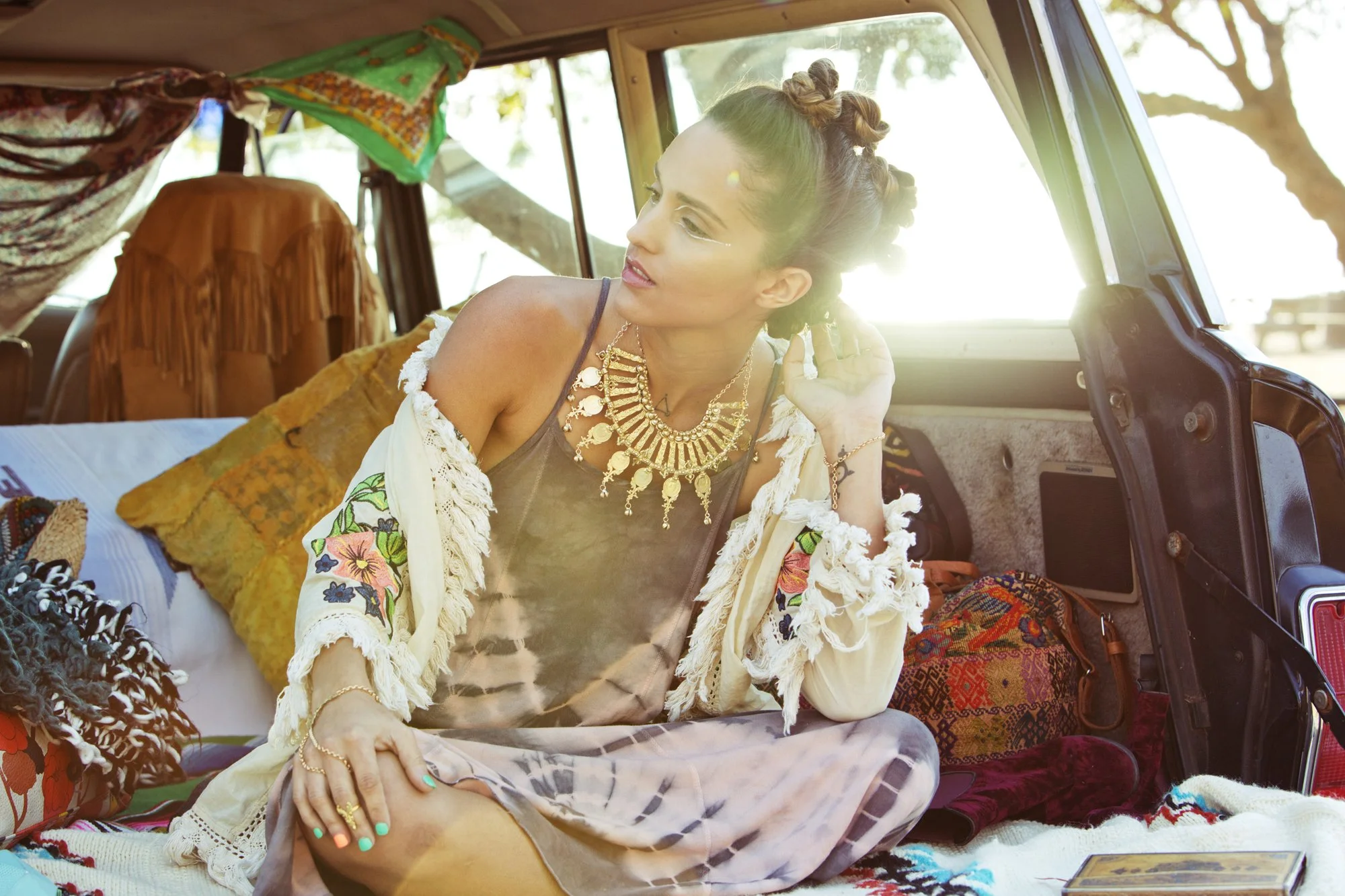 A woman with elaborate hair curls and gold jewelry sitting inside a vintage van filled with colorful blankets and pillows, with sunlight streaming through the window.