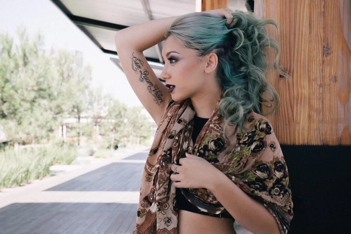 A woman with styled blonde and teal hair, dark lipstick, and tattoos, standing outdoors near a wooden wall, holding her hair up with one hand and adjusting her scarf with the other.