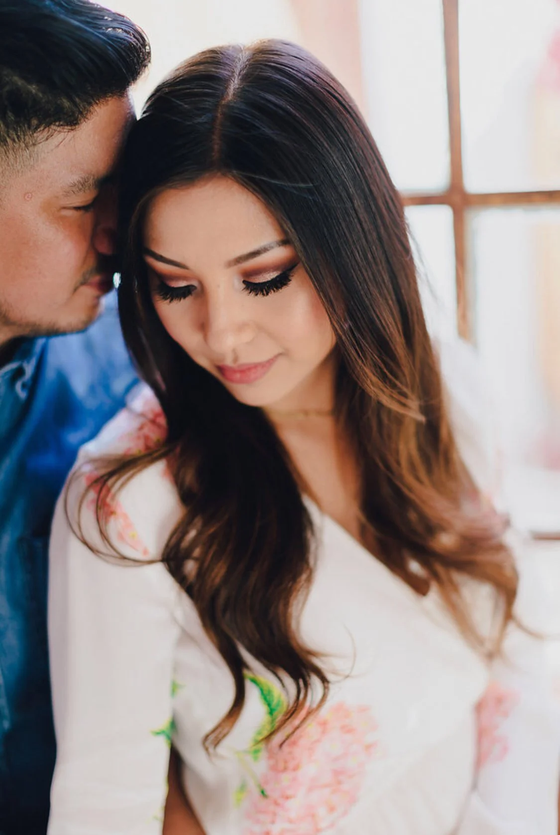 A close-up of a man and woman with their heads touching, eyes closed, inside a bright room with a window in the background.