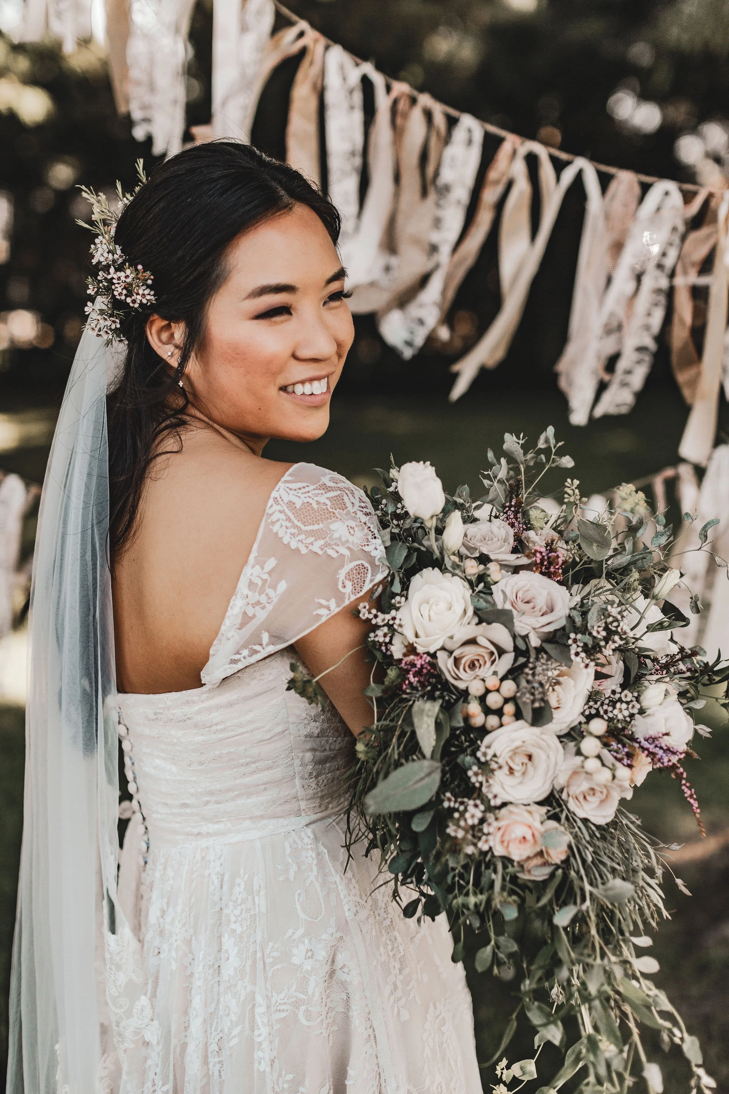 A smiling bride with dark hair styled in loose waves, wearing a lace wedding dress with floral embroidery and a sheer lace back. She has a floral hairpiece with white and purple flowers, a veil, and earrings. She is holding a large bouquet of white, 
