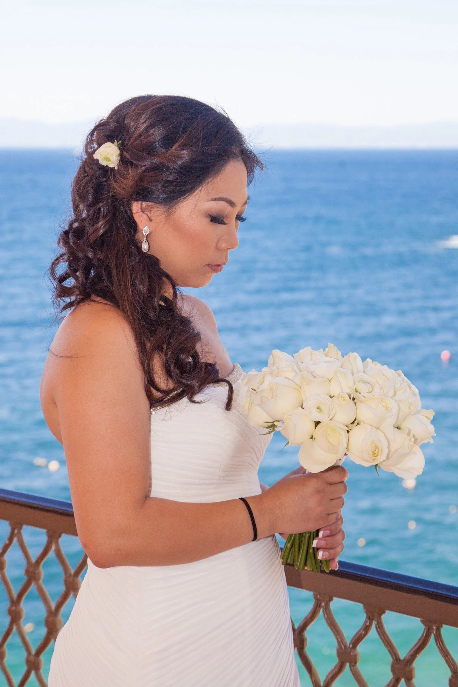 A bride with dark wavy hair adorned with a white flower, wearing jewelry, holding a bouquet of white roses, standing on a balcony overlooking the ocean.