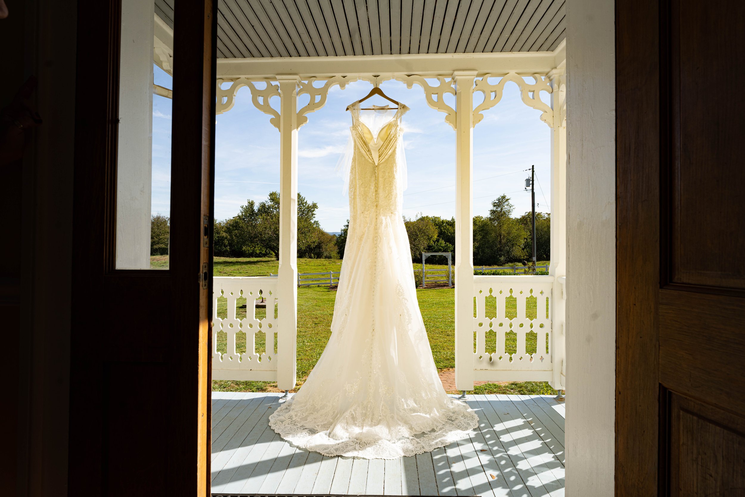 A white wedding dress hanging on a hanger outside on a porch, framed by the doorway, with a grassy yard, trees, and a blue sky in the background.