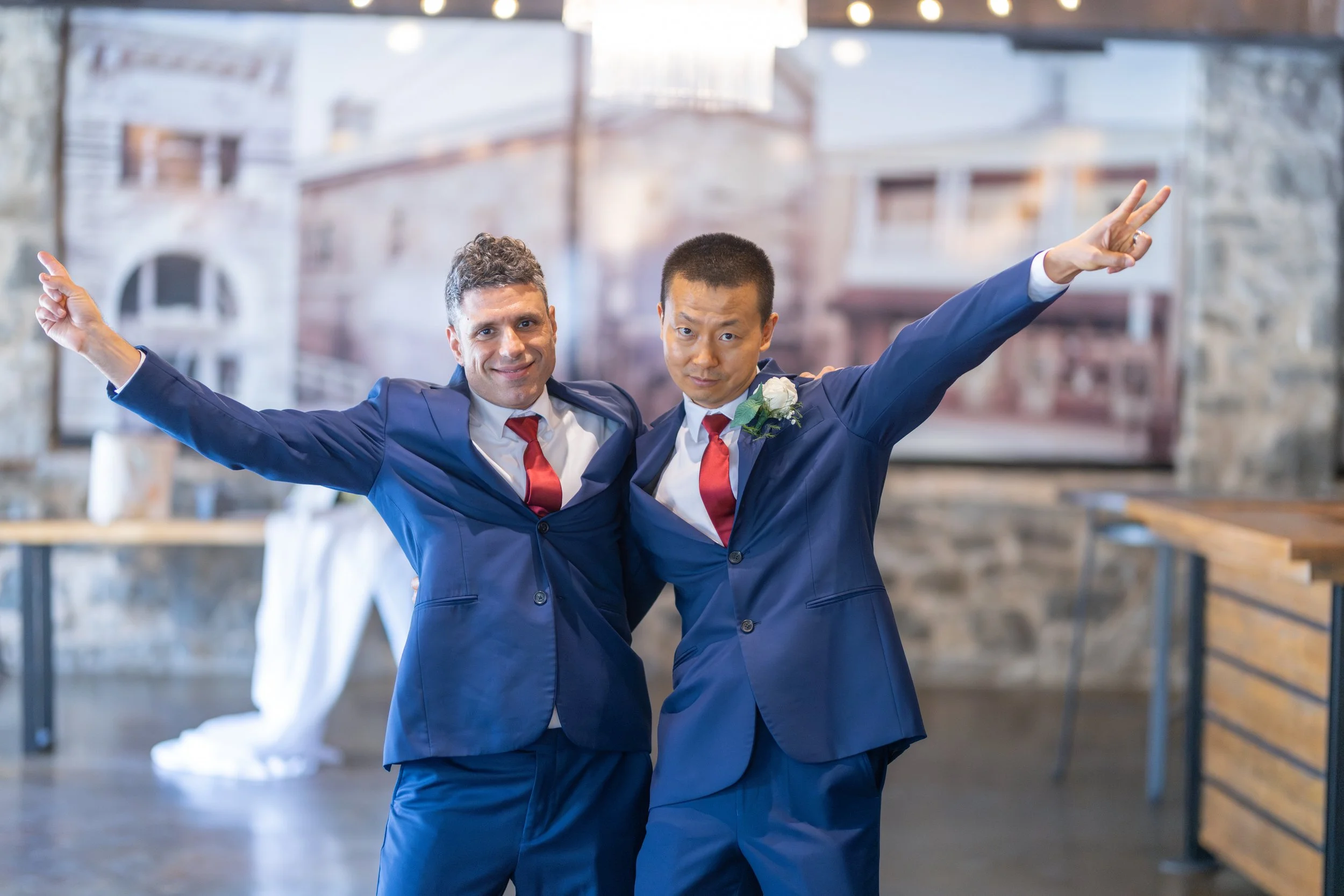 Two men in blue suits with red ties and white shirts, standing close together with arms outstretched, smiling and striking celebratory poses at an indoor event with a stone wall and artwork in the background.