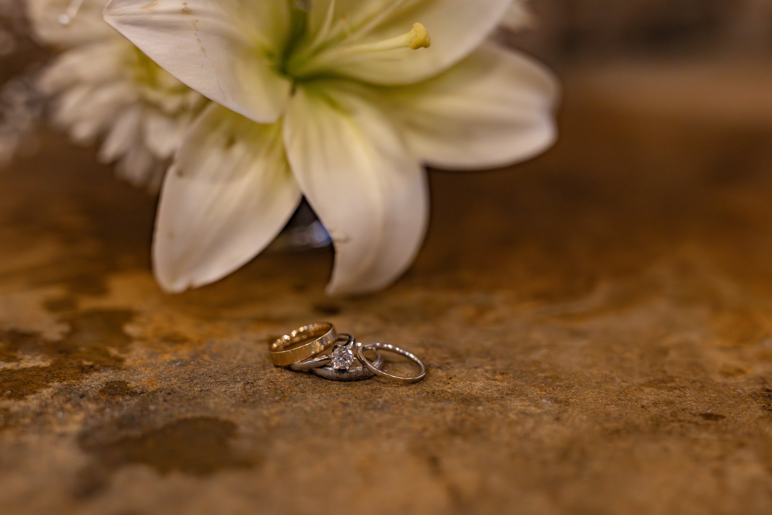 Wedding rings placed on a textured brown surface with white lilies in the background.
