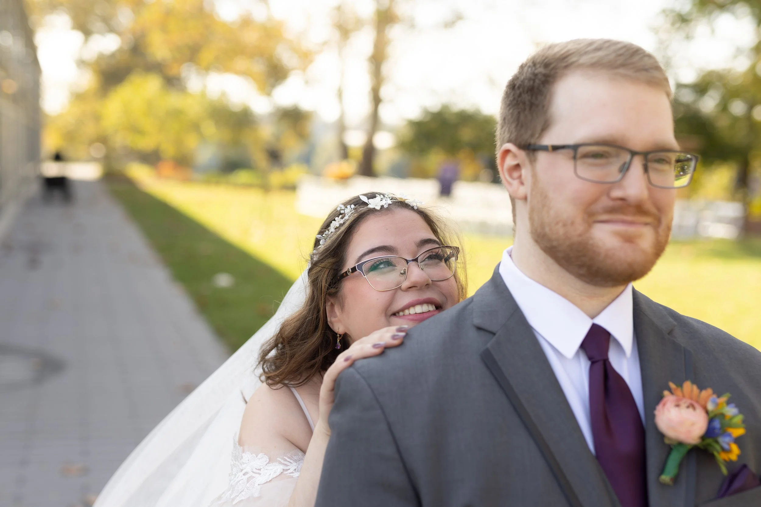 A bride and groom outdoors during daytime on a wedding day. The bride, wearing glasses, is smiling and gently touching the groom's shoulder from behind. The groom is wearing glasses, a gray suit, and a boutonniere. The background features a walkway, trees with autumn leaves, and a soft sunlight glow.