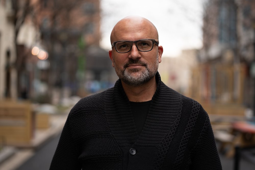 A middle-aged man with a bald head, glasses, and a beard standing outdoors in an urban setting with trees and buildings in the background.