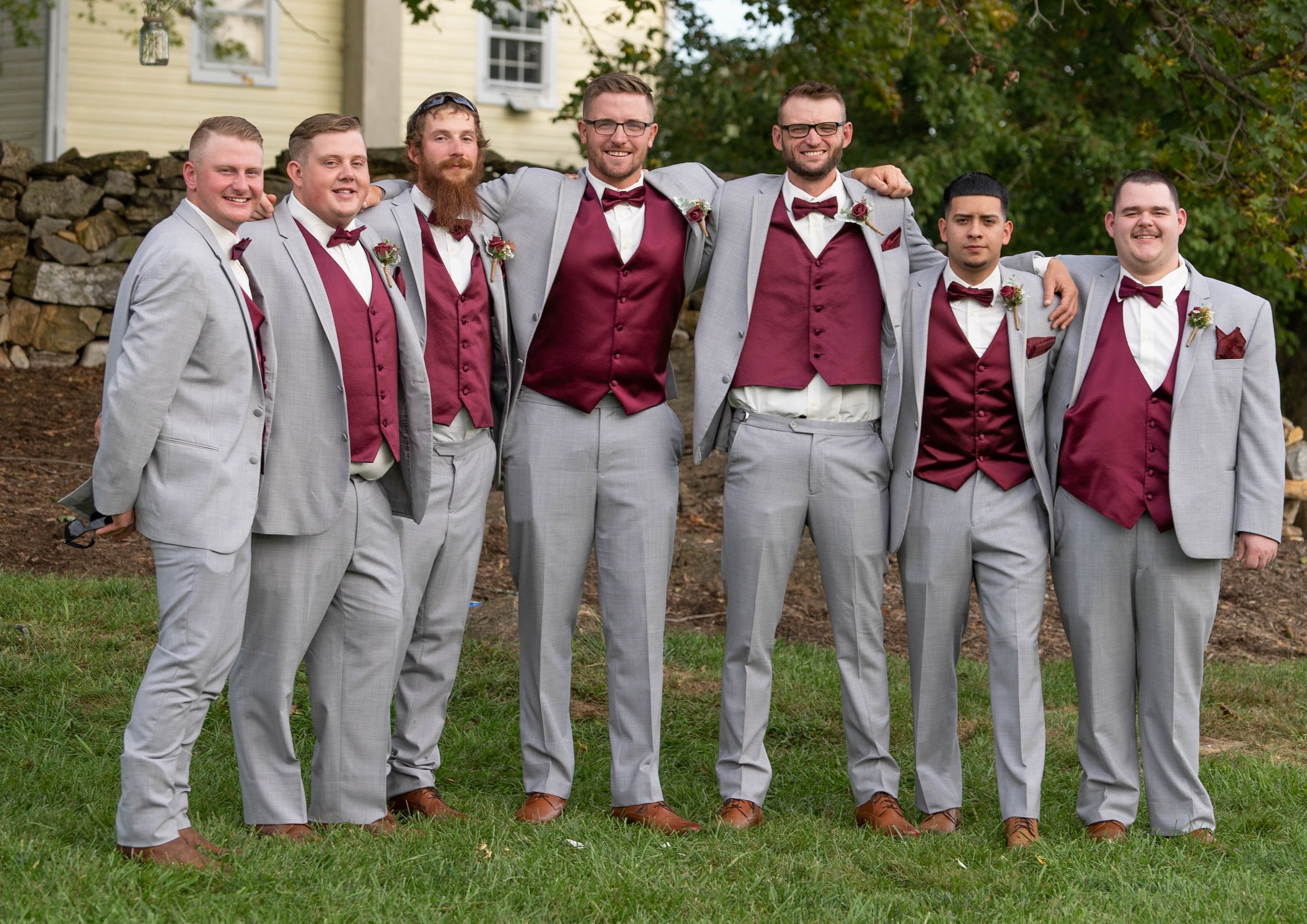 Group of eight men in matching suits standing outdoors, smiling, with arms around each other's shoulders, on grass with trees and a house in the background.