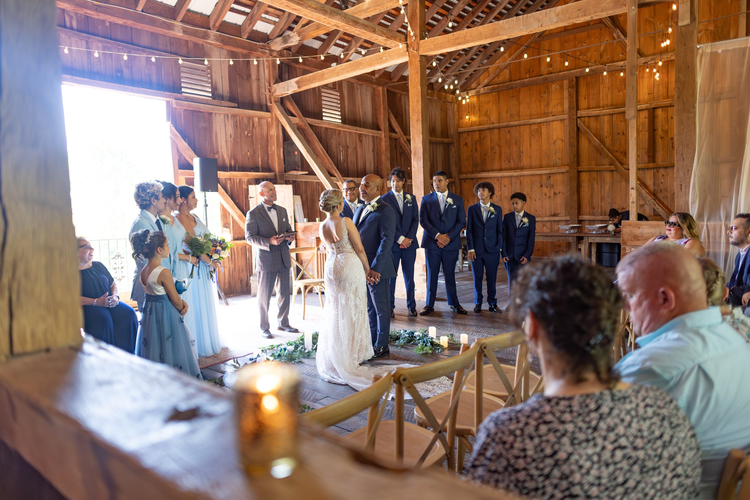 A wedding ceremony taking place in a rustic barn with wooden interior, string lights, and natural sunlight, featuring a bride and groom exchanging vows surrounded by their wedding party and guests.