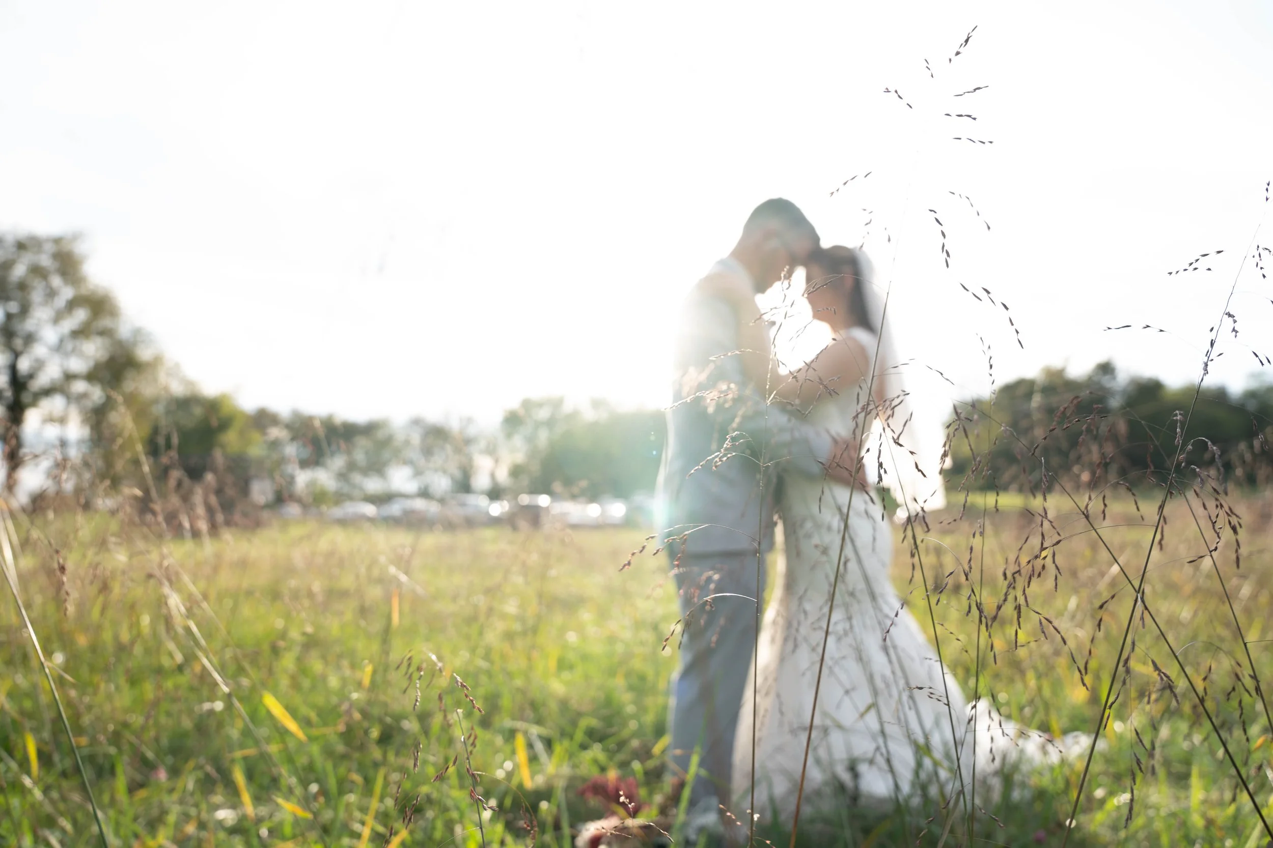 A couple in wedding attire embracing in a grassy field with sunlight creating a backlit effect