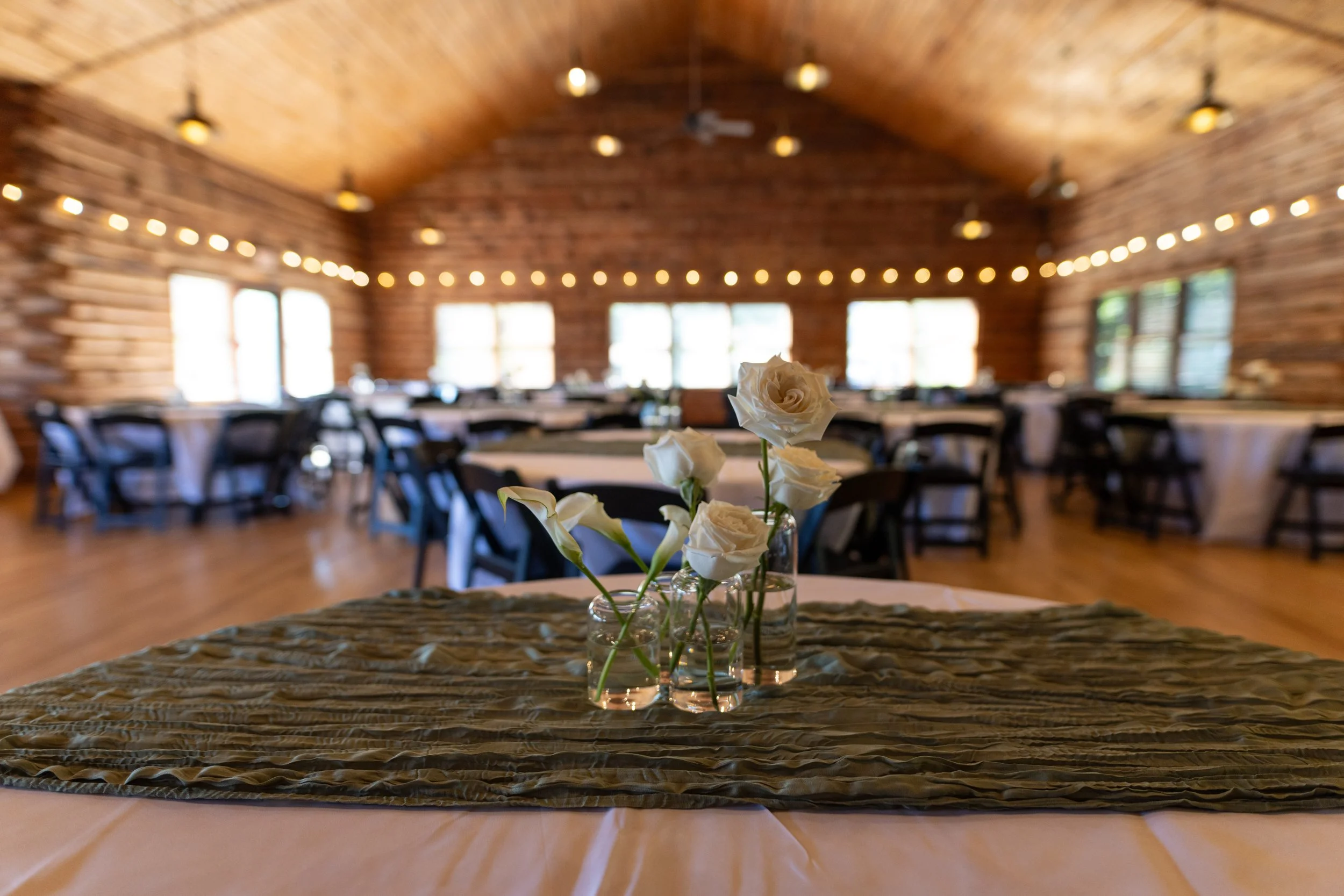 A table with white roses in small glass vases on a textured green table runner, set in a rustic wooden banquet hall with string lights overhead.