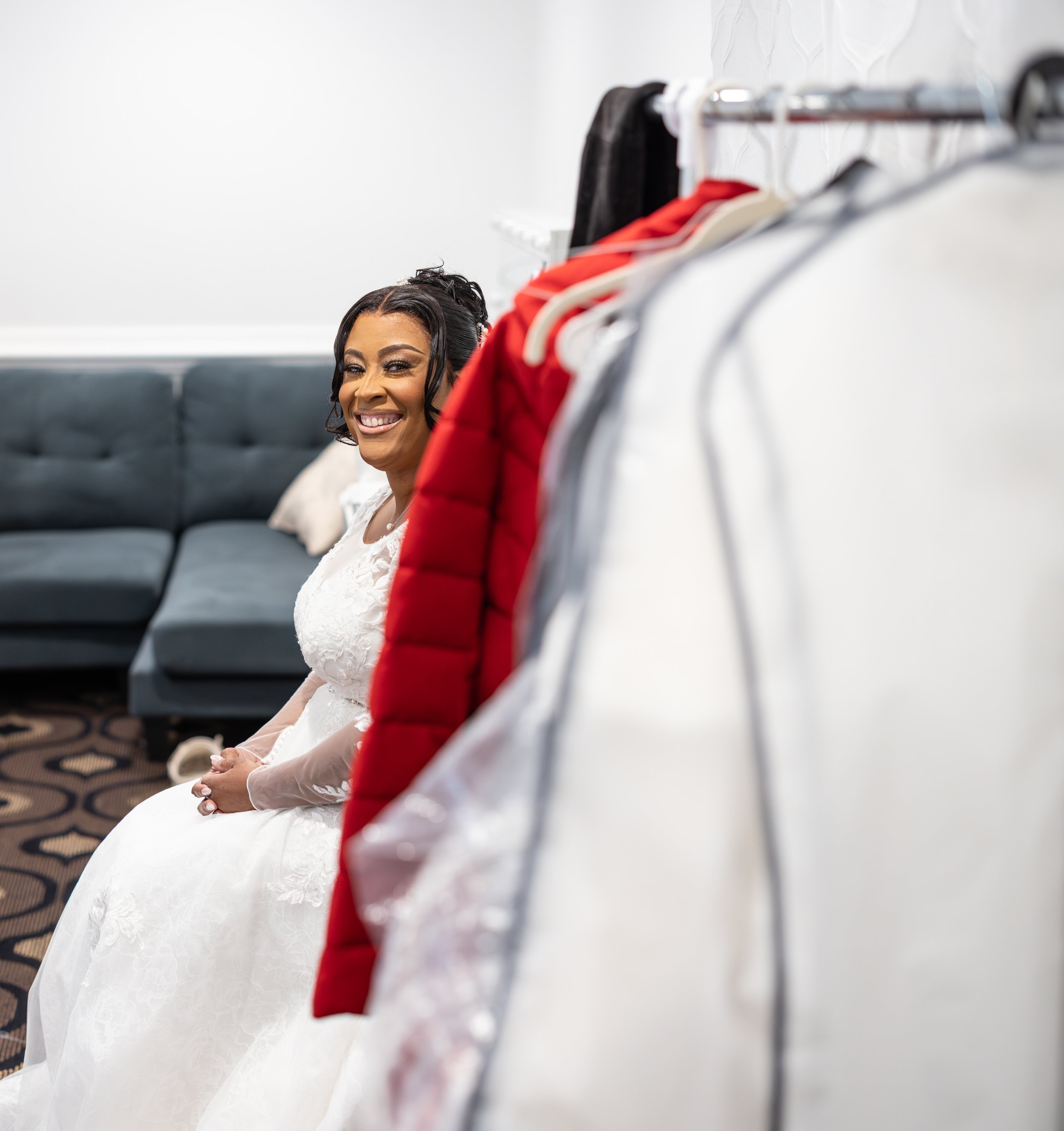 A bride in a white wedding gown sitting on a couch, smiling, with a clothing rack of colorful jackets and shirts in the foreground.