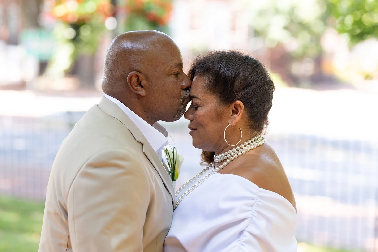 A mature couple sharing a wedding kiss outdoors. The man in a beige suit and the woman in a white dress with pearl jewelry. The background includes blurred trees and sunlight.