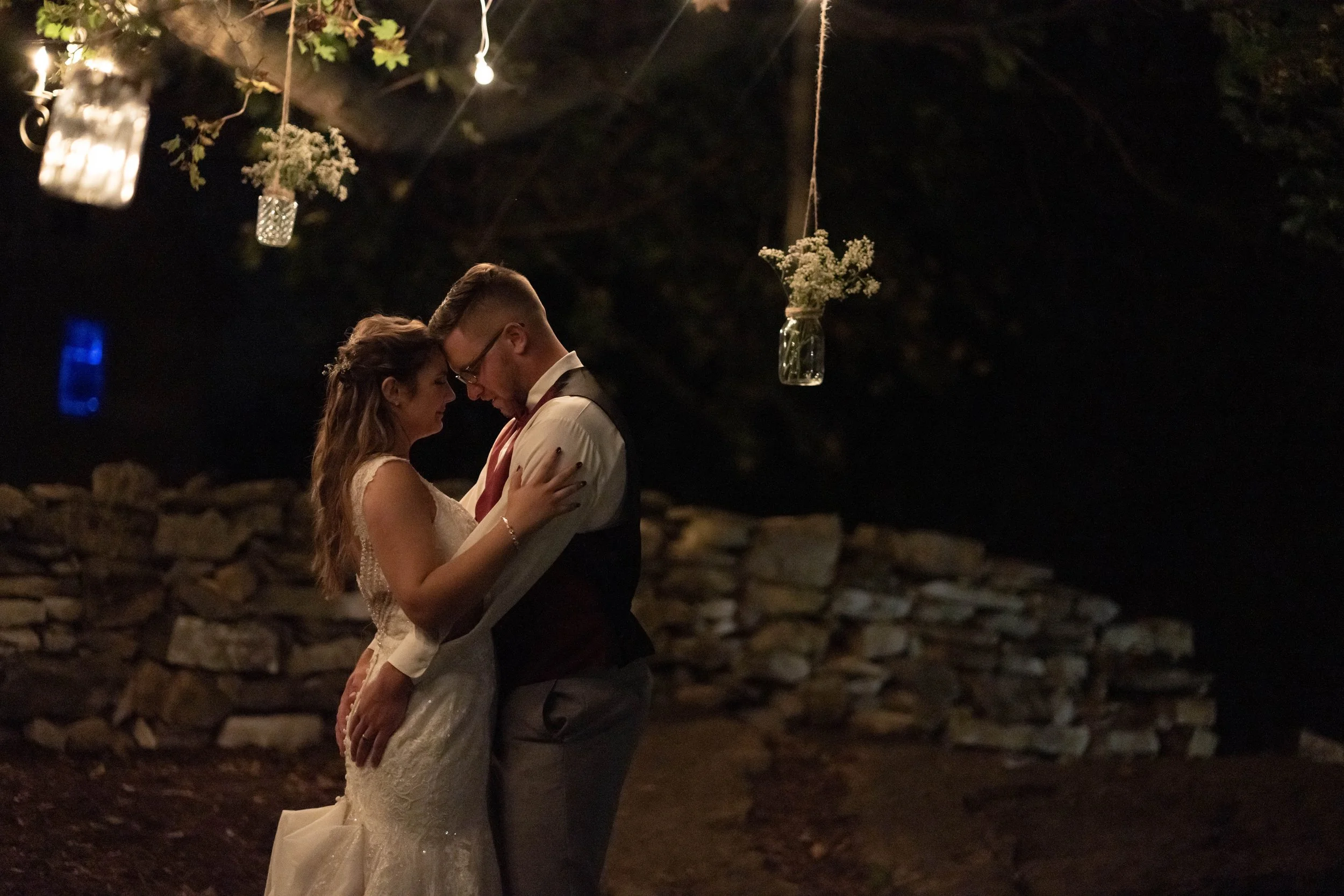 A bride and groom share an intimate dance outdoors at night, with hanging mason jars and flowers above them and a stone wall in the background.