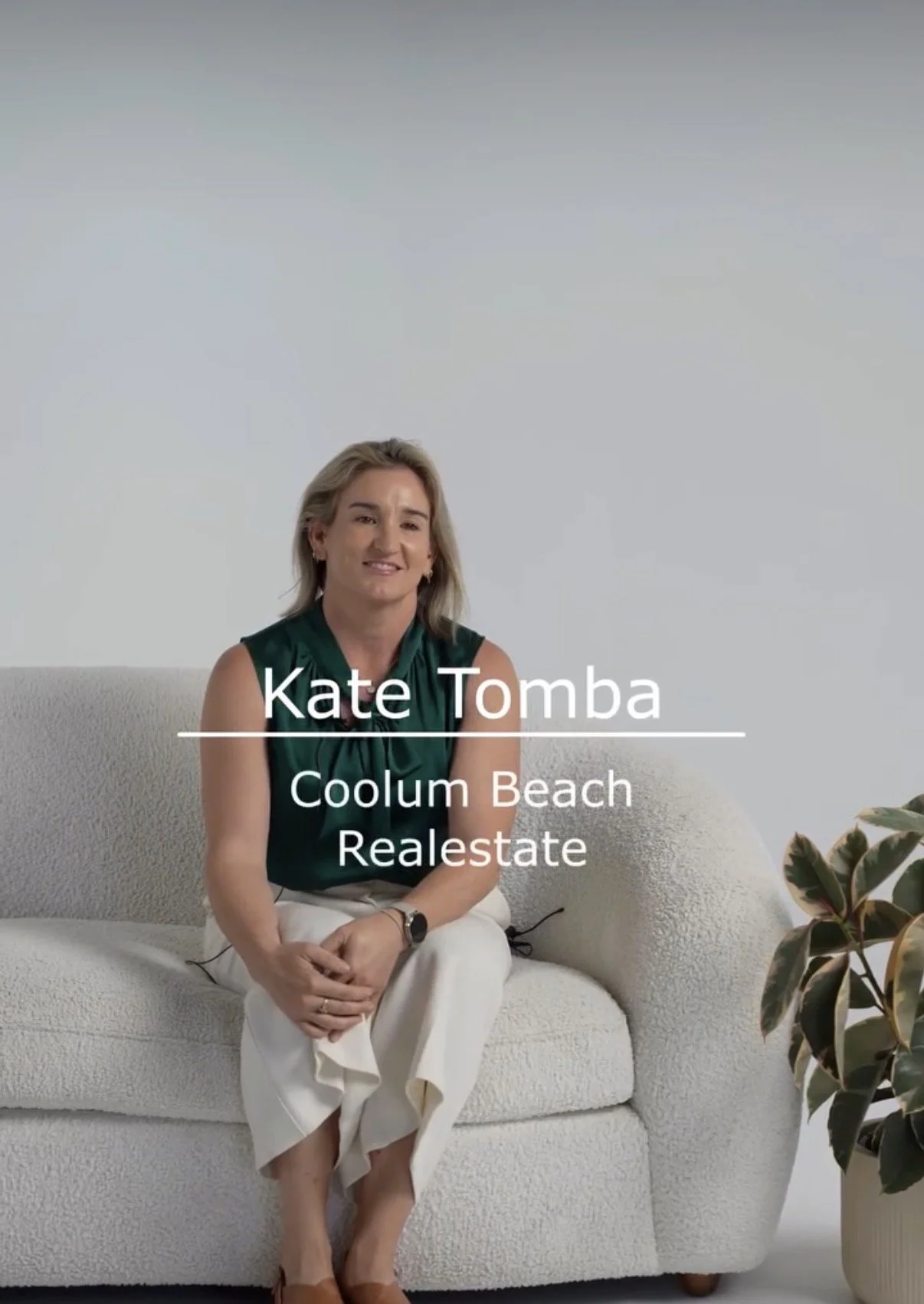 A woman sitting on a white textured sofa in a minimalistic, modern room. She wears a dark green sleeveless top and white pants. She is smiling slightly, with a plant visible in the lower right corner. Text overlay reads: 'Kate Tomba, Coolum Beach, Realestate.'