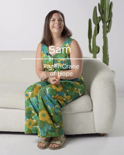 Woman smiling and sitting on a white sofa, wearing a colorful, floral dress, with a tall cactus plant in the background.