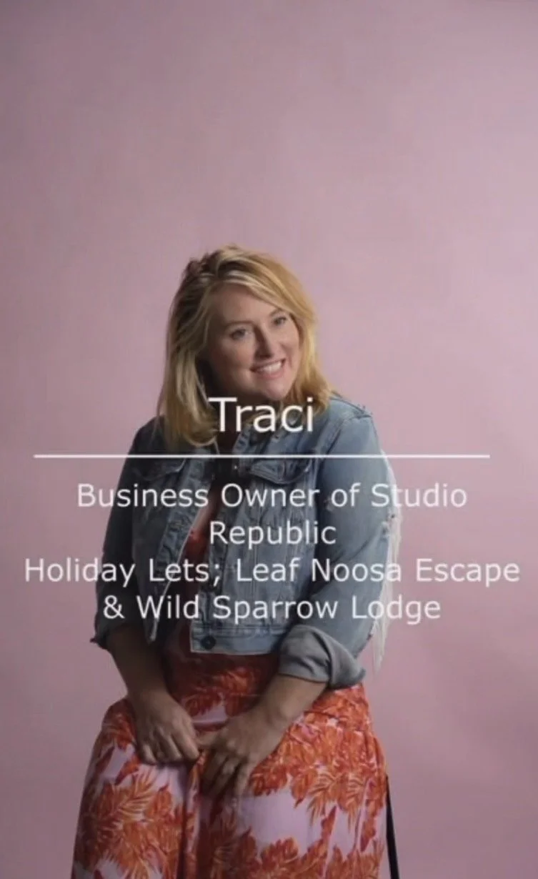 A woman with shoulder-length blonde hair, wearing a denim jacket and orange floral pants, smiling while sitting against a pink background. The overlay text reads: Traci, Business Owner of Studio Republic, Holiday Lets; Leaf Noosa Escape & Wild Sparrow Lodge.