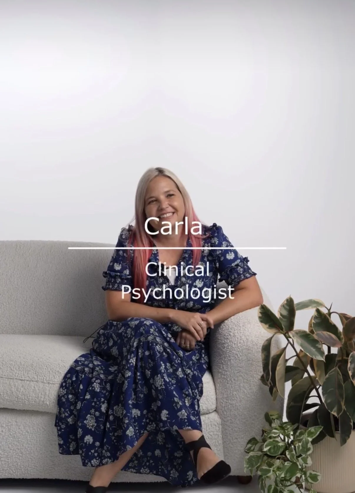 Smiling woman with blonde and pink hair sitting on a beige couch, wearing a blue floral dress, next to a potted plant, with the text "Carla, Clinical Psychologist" overlayed.
