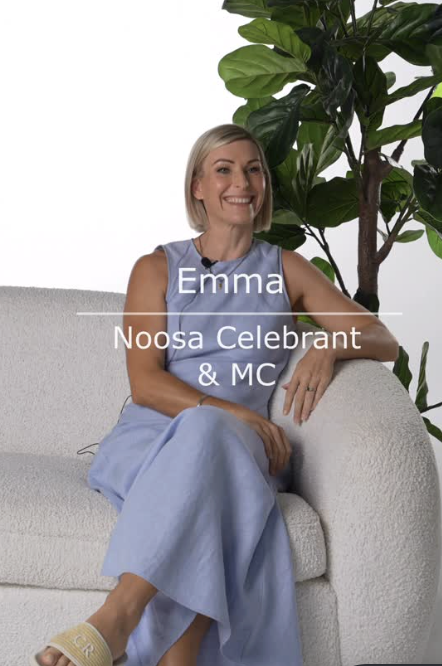 A woman with short blonde hair, sitting on a light-colored sofa, smiling, wearing a sleeveless lavender top and light blue pants, with a large green leafy plant behind her.