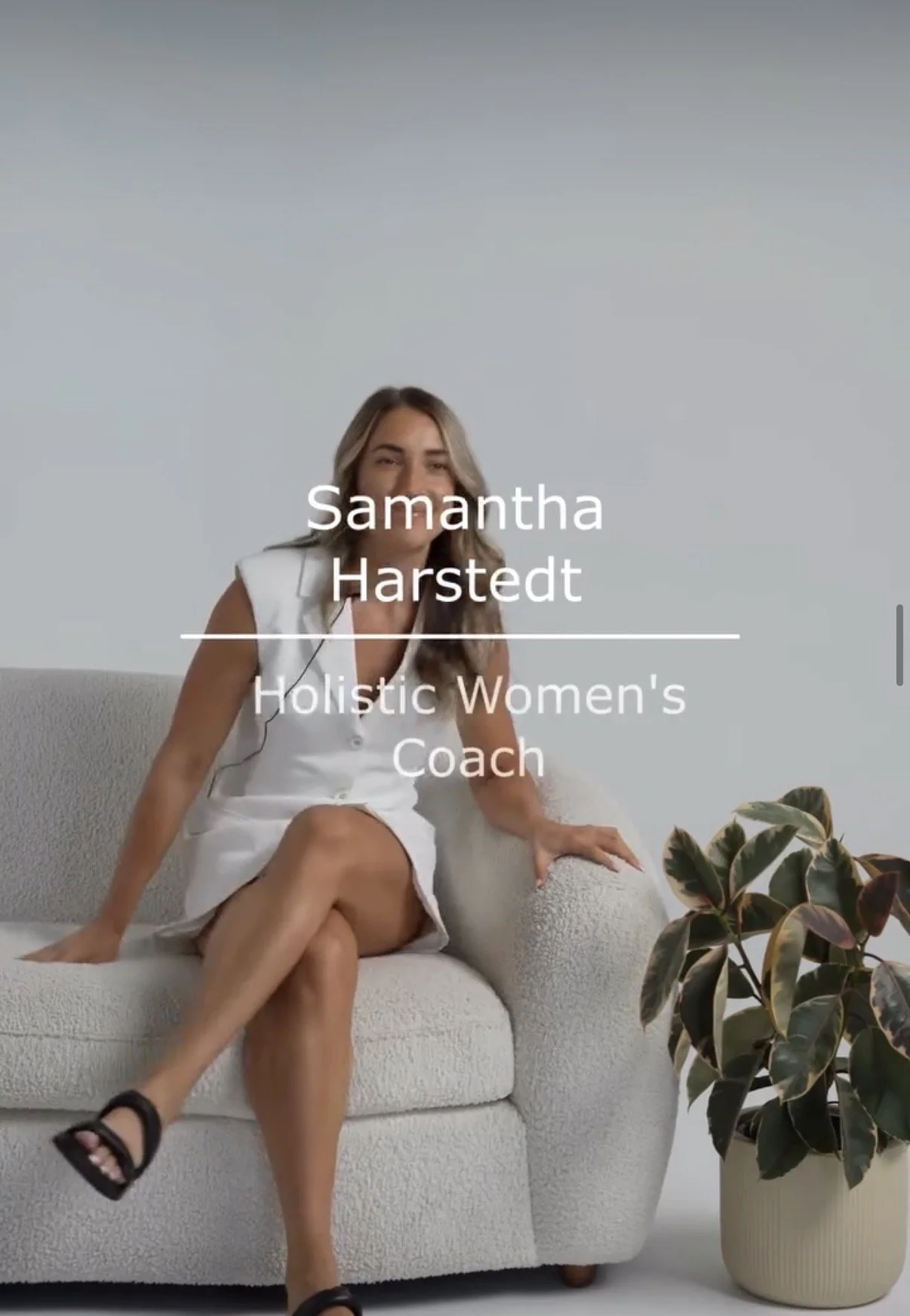 A woman sitting on a white textured sofa with her legs crossed, wearing a white sleeveless dress and black sandals. There is a potted plant with dark green and purple leaves on the floor beside her. The background is a plain white wall, and the woman's name, Samantha Hartsedt, is overlaid along with the title "Holistic Women's Coach".