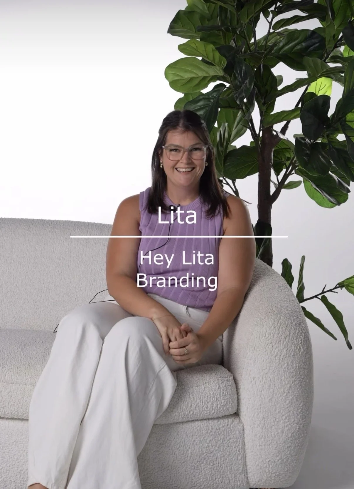 A woman sitting on a white textured sofa with a large green leafy plant behind her, smiling and looking at the camera. She is wearing glasses, a sleeveless lavender top, and white pants. The image has a white background with text overlay that reads: "Lita" and "Hey Lita Branding."