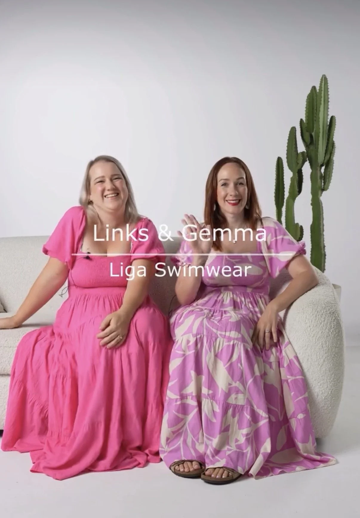 Two women sitting on a white sofa, smiling and waving. One is wearing a bright pink dress, and the other a pink and purple patterned dress. There is a large cactus plant behind them. Text overlay reads "Links & Gemma Liga Swimwear."