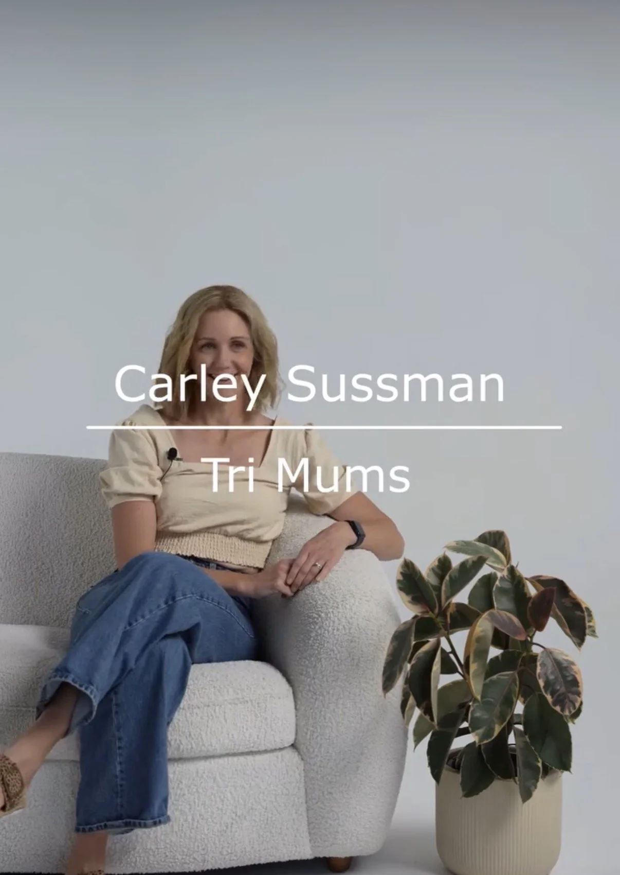 A woman sitting on a white textured sofa with a plant in a large white pot on the right. She has blonde hair, is wearing a light beige top and blue jeans, and is smiling. Text overlay reads 'Carley Sussman' and 'Tri Mums.'