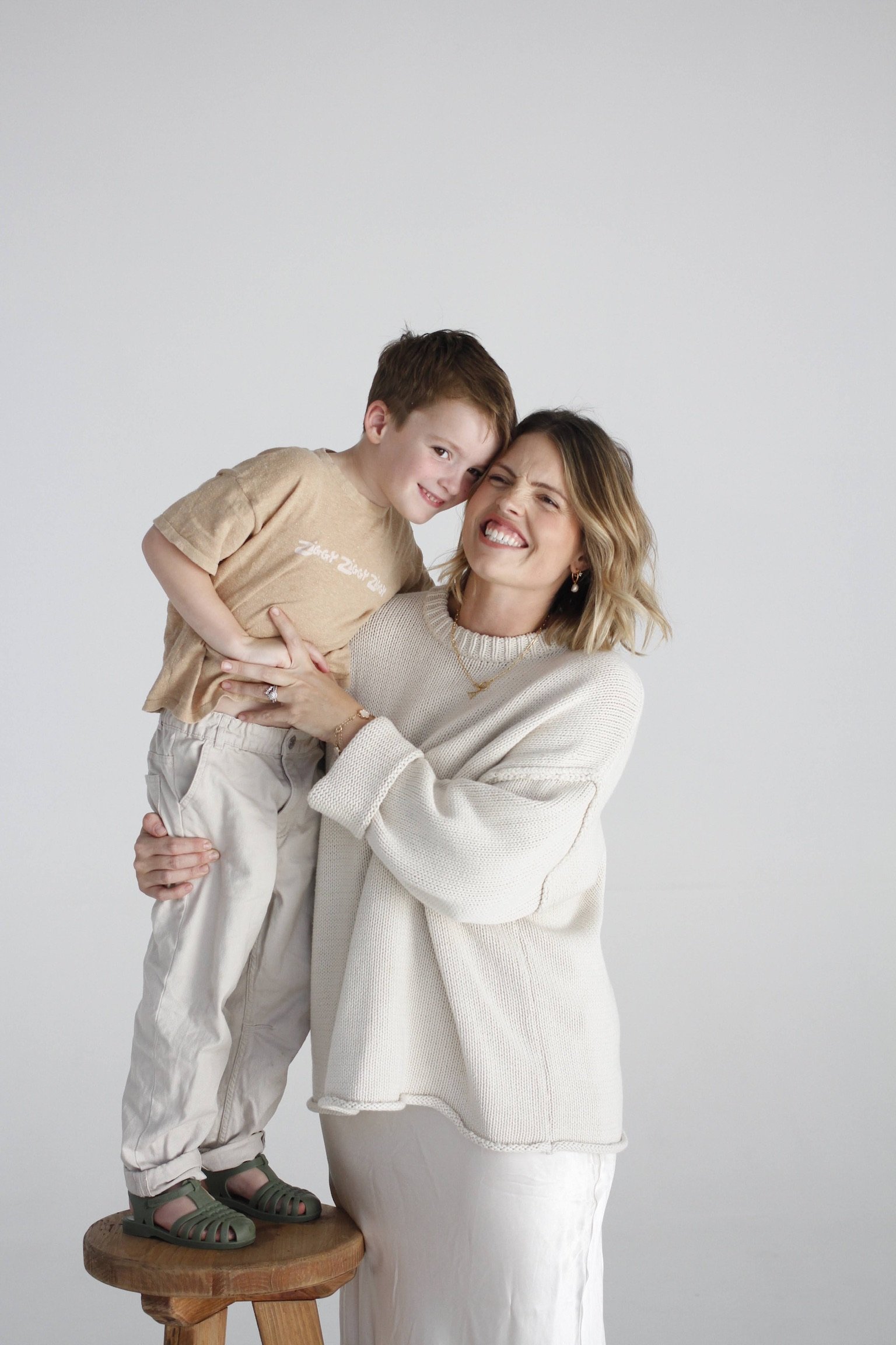 A woman with blonde wavy hair smiling and holding a young boy with short brown hair, who is smiling and leaning his head on her face. The woman is wearing a beige sweater, and the boy is wearing a tan T-shirt and beige pants. The boy is standing on a round wooden stool, and he is wearing green sandals. The background is a plain white wall.