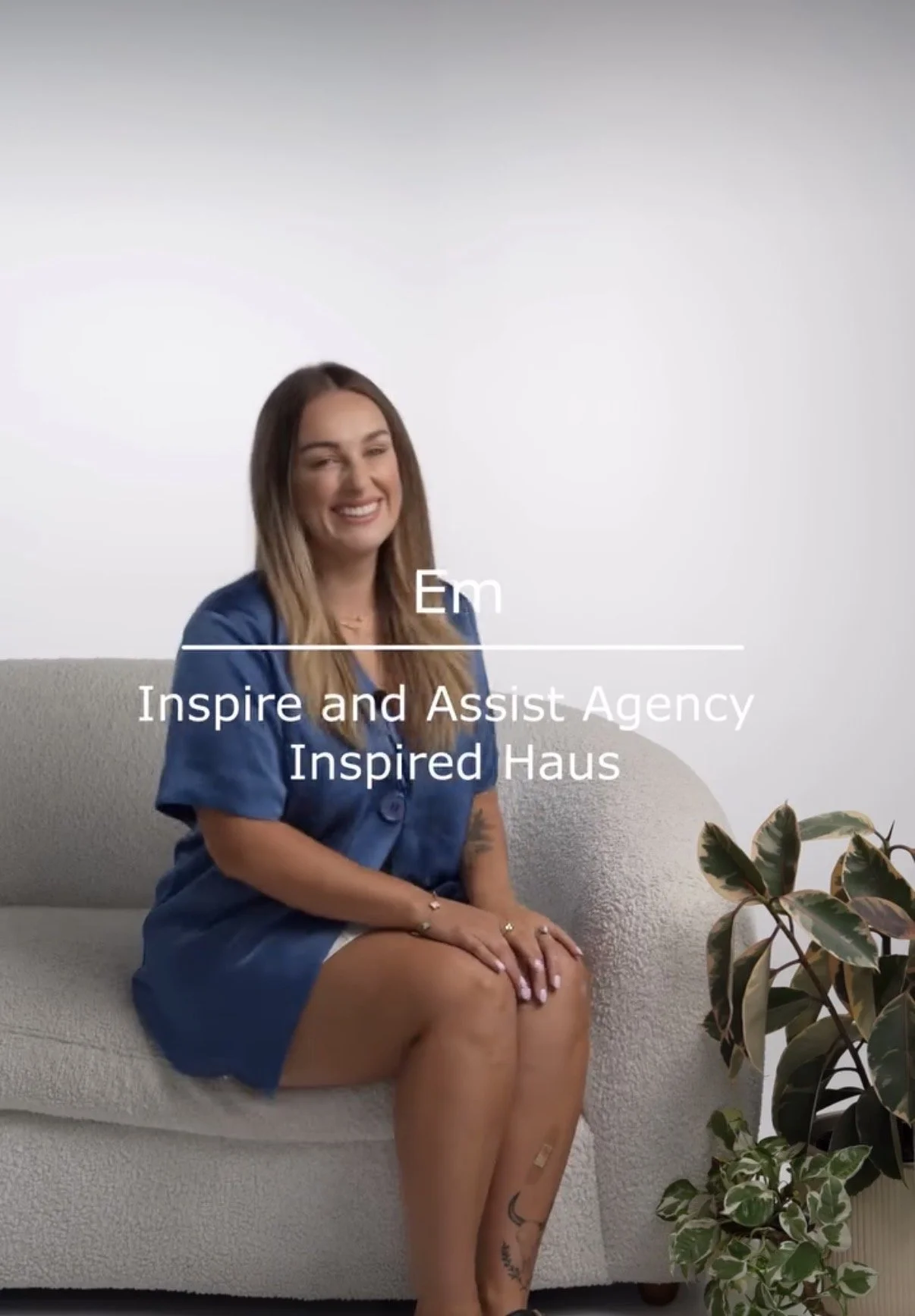 Smiling woman sitting on a gray couch with a potted plant nearby, wearing a blue dress, with text overlay that reads 'Em' and 'Inspire and Assist Agency Inspired Haus'.