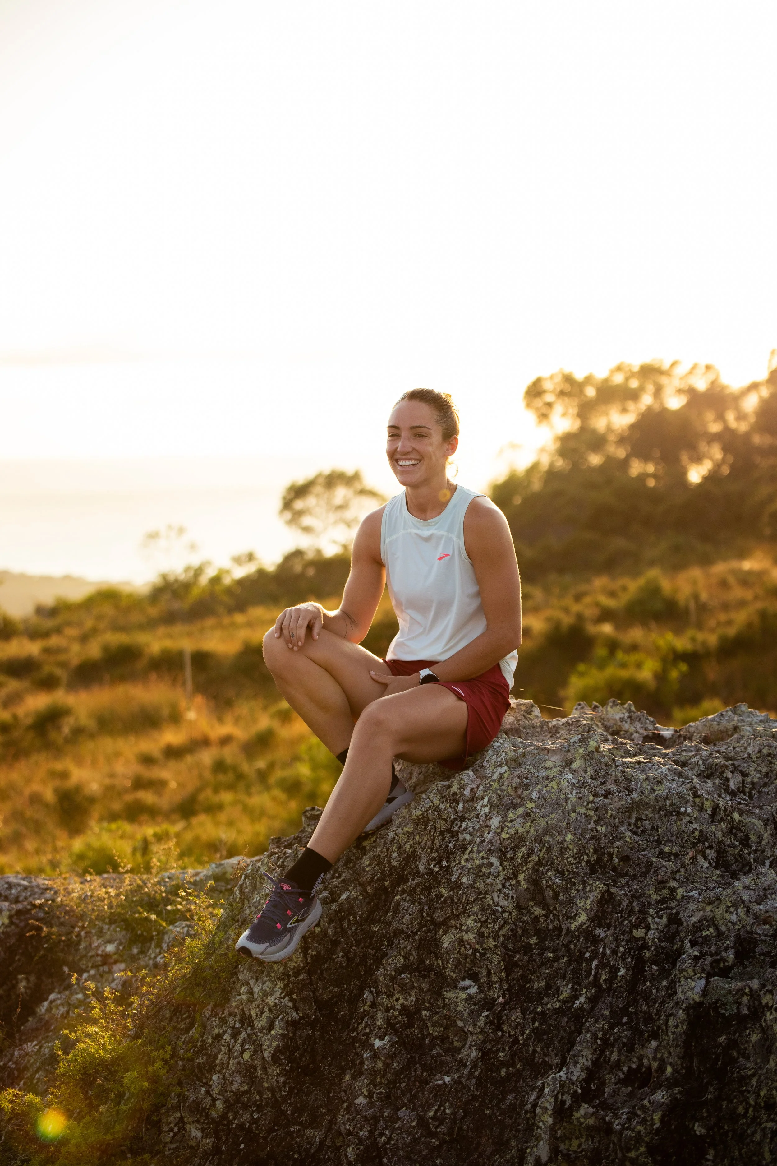 A woman with brown hair tied back, sitting on a rock outdoors at sunset, smiling, wearing a white sleeveless athletic top, red shorts, black sneakers, and black socks, with a background of trees and a golden sky.