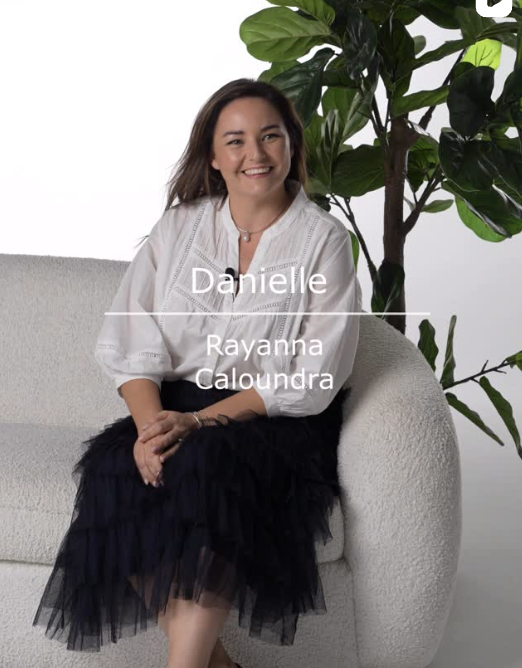A woman named Danielle sitting on a light-colored modern sofa with a big green leafy plant behind her, wearing a white blouse and a black tulle skirt, smiling at the camera.