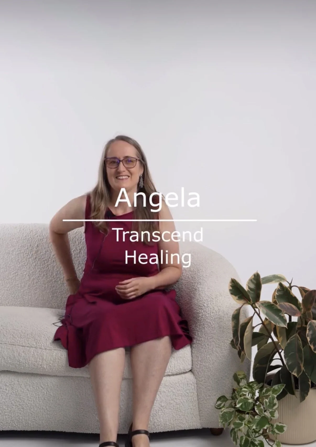 A woman with long hair and glasses sitting on a cream-colored couch, wearing a burgundy dress, smiling, with a houseplant nearby. Text on the image reads: Angela, Transcend Healing.