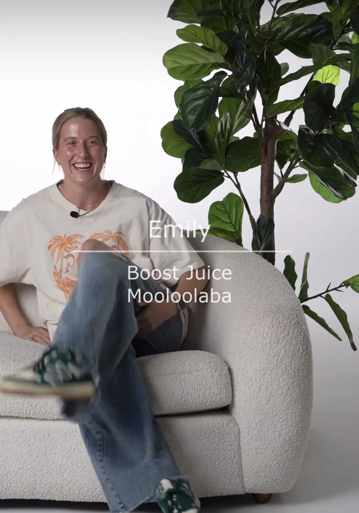 A woman smiling and sitting on a beige textured sofa next to a large green houseplant. Text on the image reads: 'Emily, Boost Juice, Mooloolaba.'