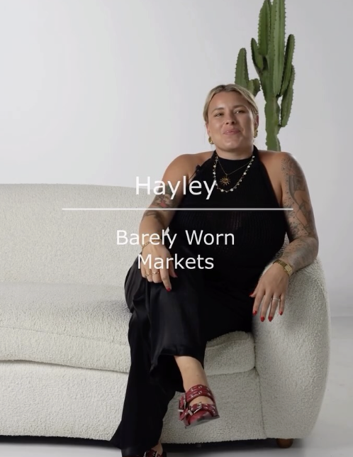 A woman with tattoos sits on a white sofa in front of a white background with a tall cactus plant. She wears a black sleeveless top, black pants, red sandals, and gold jewelry, and has short blonde hair.