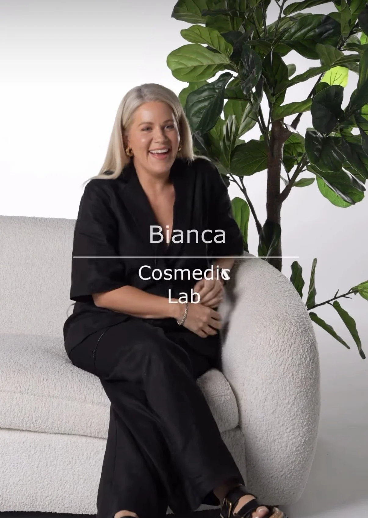 Blanca sitting on a white textured sofa with a large green leafy plant behind her, smiling. She is wearing a black outfit and gold earrings, with her right arm resting on her lap. The text overlay says 'Blanca Cosmedic Lab'.