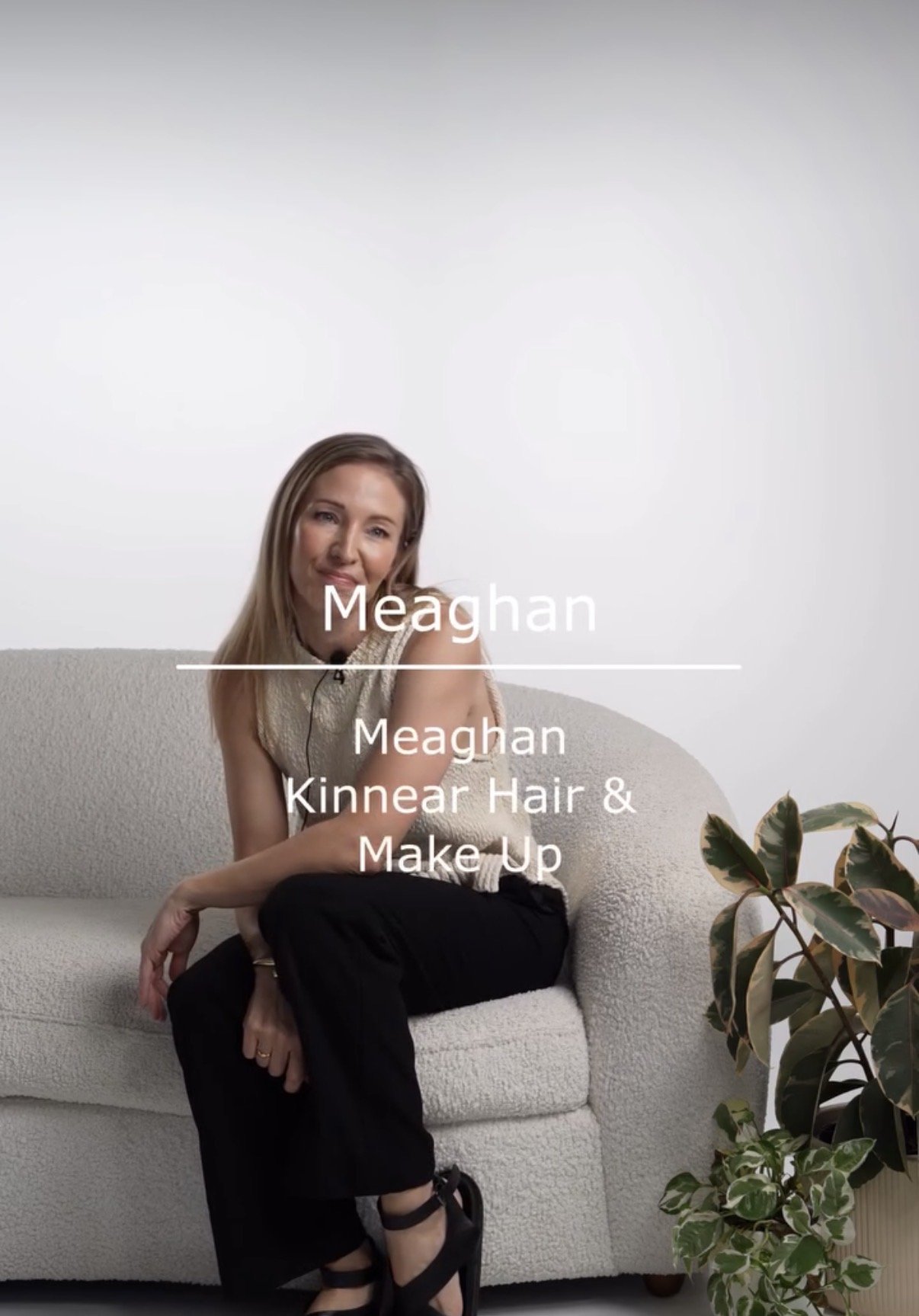 A woman sitting on a white couch with a beige cushion, wearing a sleeveless beige top and black pants. She has long hair and is smiling, with a plant visible on the right side of the image. The text overlay reads 'Meaghan - Meaghan Kinnear Hair & Make Up'.