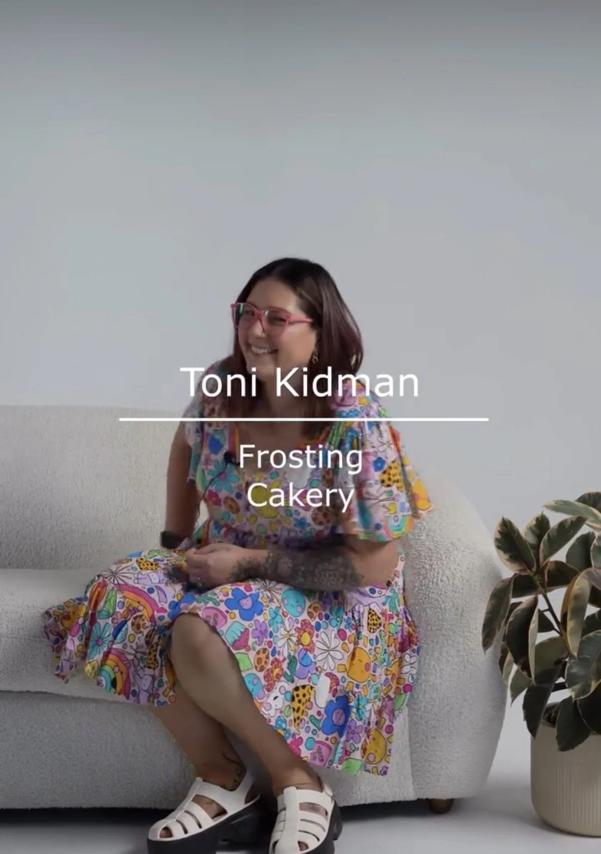 A woman with glasses and long hair, wearing a colorful, patterned dress, sitting on a white sofa with her legs crossed. There is a potted plant on her right side.