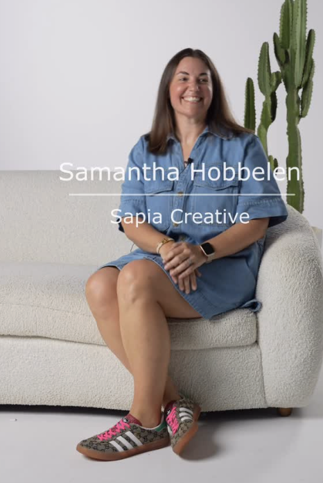 A woman sitting on a white couch with a cactus plant behind her, wearing a denim dress and sneakers, smiling at the camera.