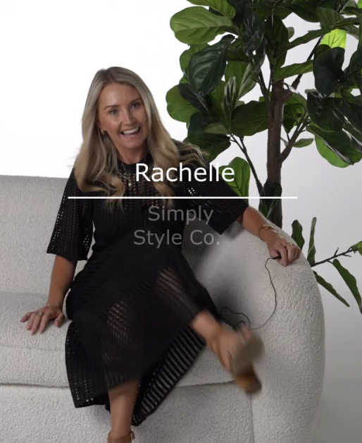 Woman sitting on a gray sofa next to a large green leafy plant, smiling and wearing a black dress with cutout details.