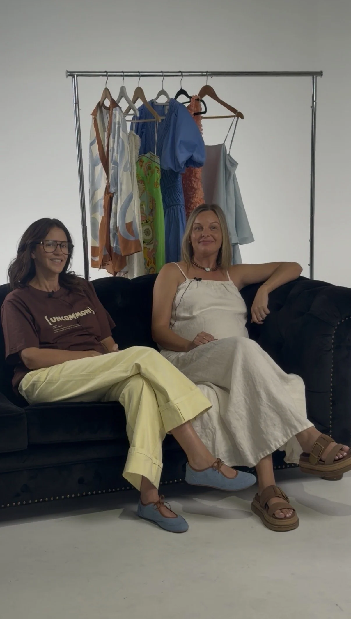 Two women sitting on a black couch with a clothing rack filled with colorful dresses behind them. One woman wears yellow pants and blue shoes, the other wears a white dress and tan sandals.