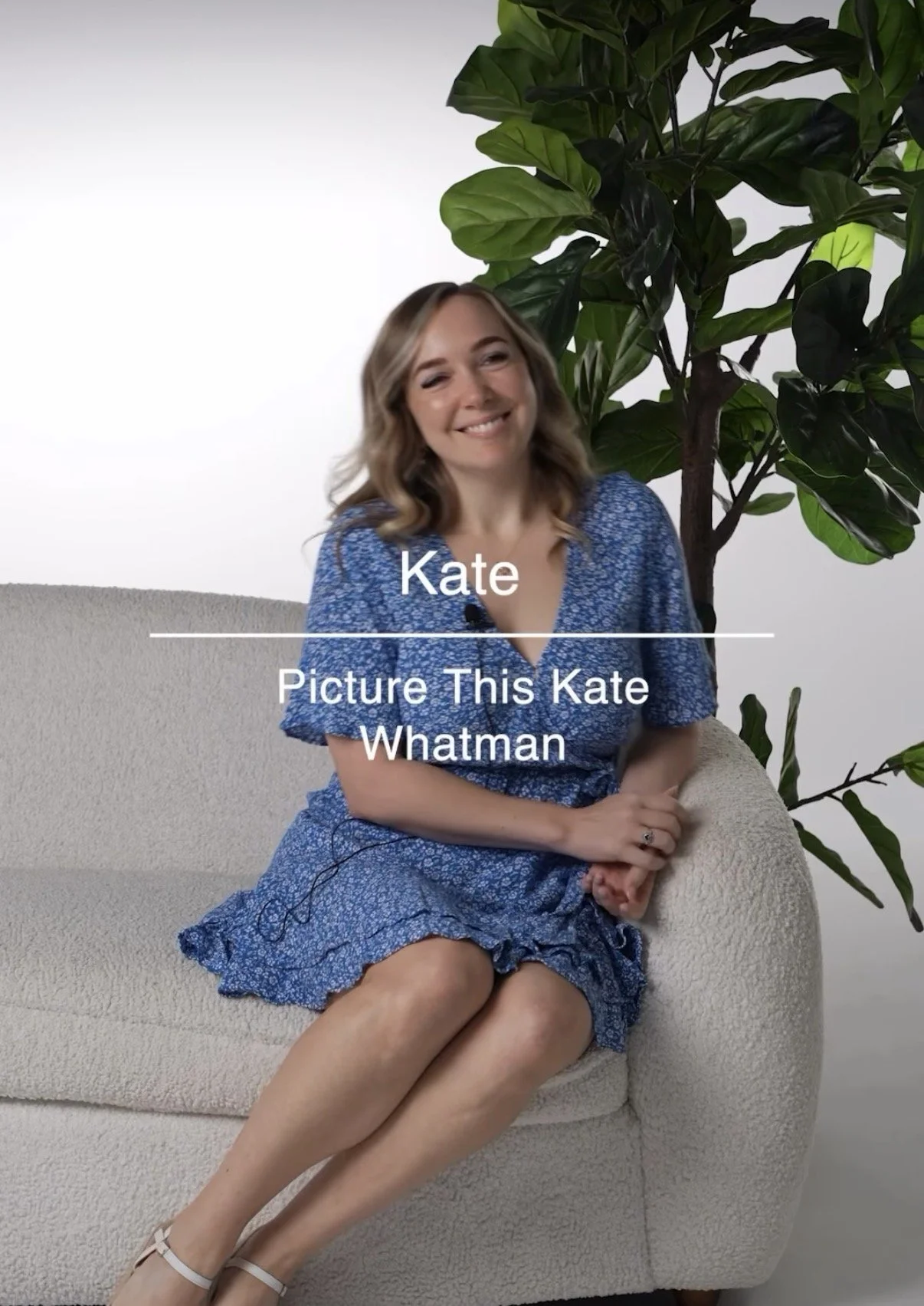 A woman with shoulder-length wavy hair smiling, wearing a blue patterned dress, sitting on a beige couch in front of a white wall with a large leafy green plant nearby.