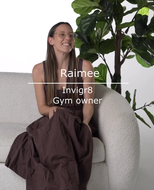 A woman with long brown hair and glasses sitting on a light-colored sofa next to a large green plant. She is smiling and wearing a dark sleeveless top and a long brown skirt. Text on the image reads: 'Raimee, Invigr8, Gym owner'.