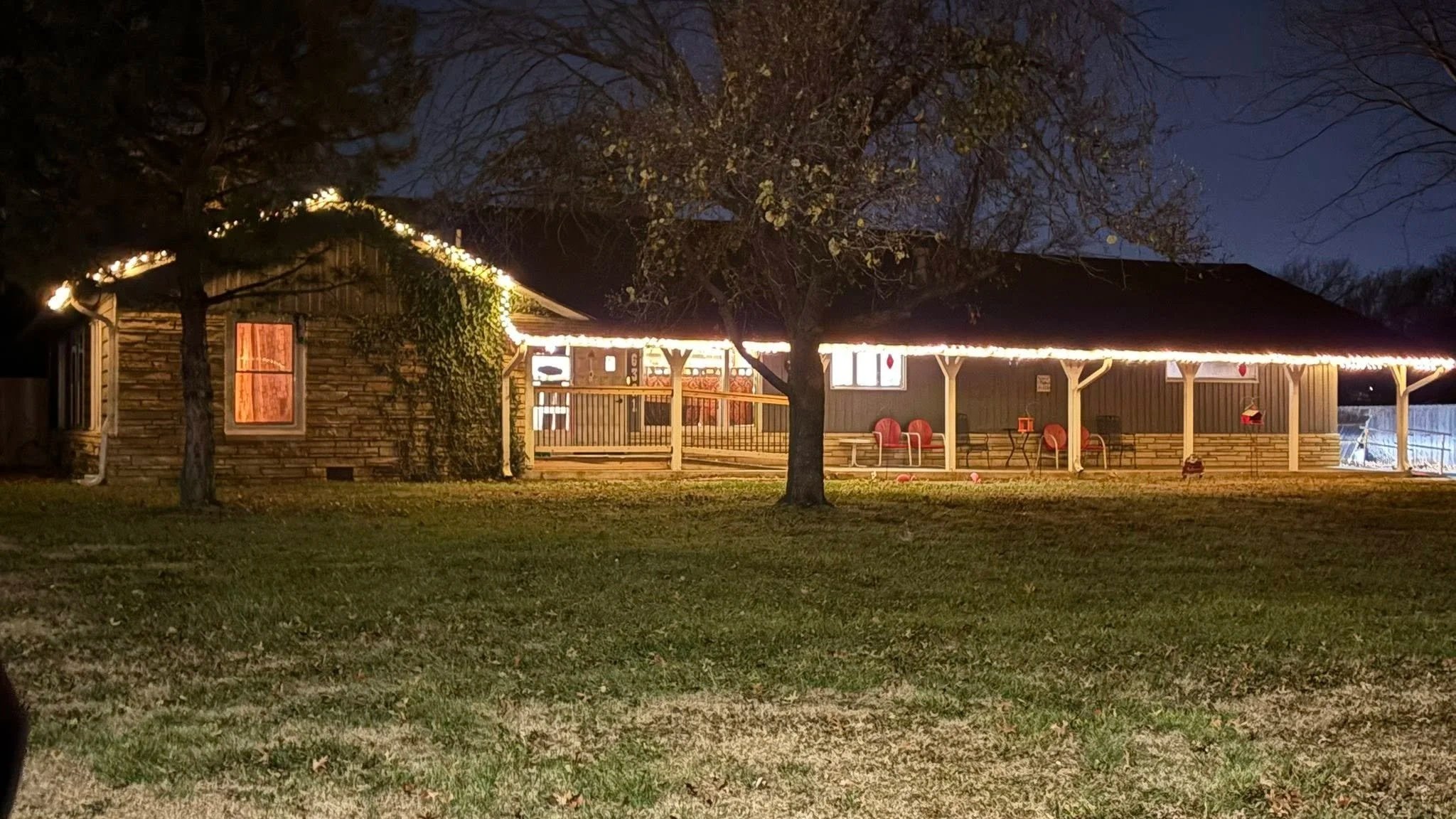 Nighttime view of a house decorated with string lights along the roofline, with a covered porch featuring red chairs, set in a yard with trees and grass.