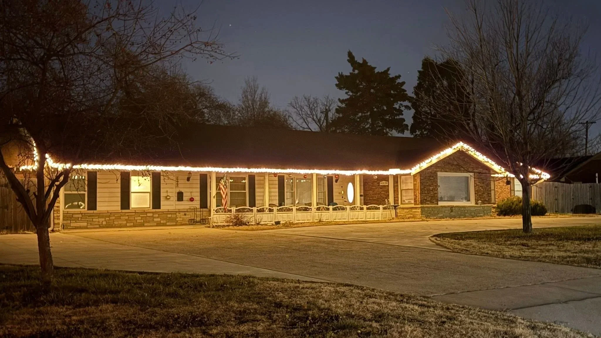A single-story house decorated with holiday string lights on the roofline, set at night. The house has beige siding with brick accents, black shutters, and a small porch with a white railing. There are bare trees in the front yard, and the driveway is visible in the foreground.