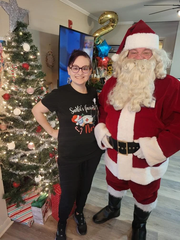 A woman standing next to Santa Claus with a Christmas tree and presents in the background.
