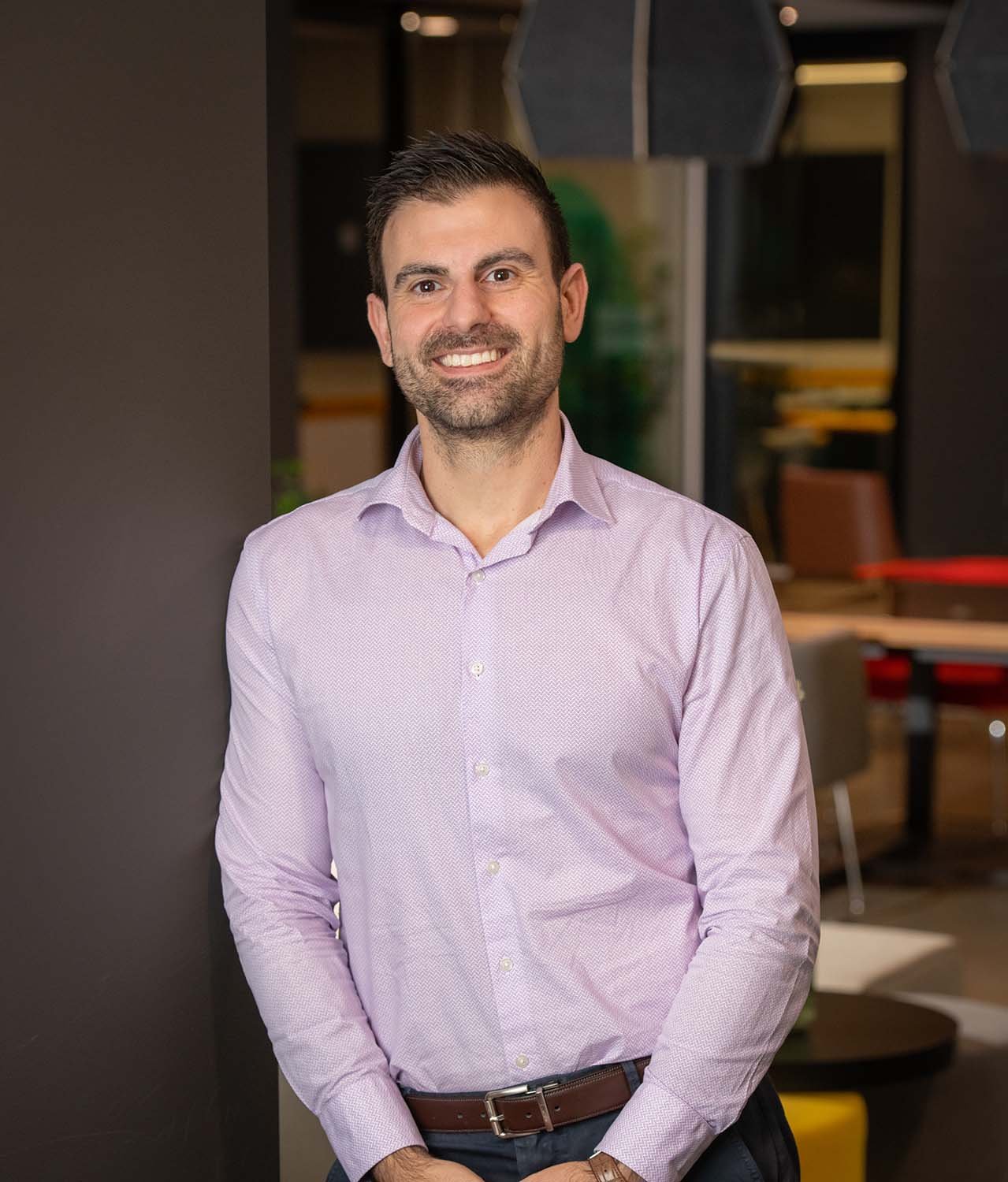 A man with short dark hair and a beard, wearing a light purple dress shirt, smiling while standing indoors with a dark wall and blurred background.