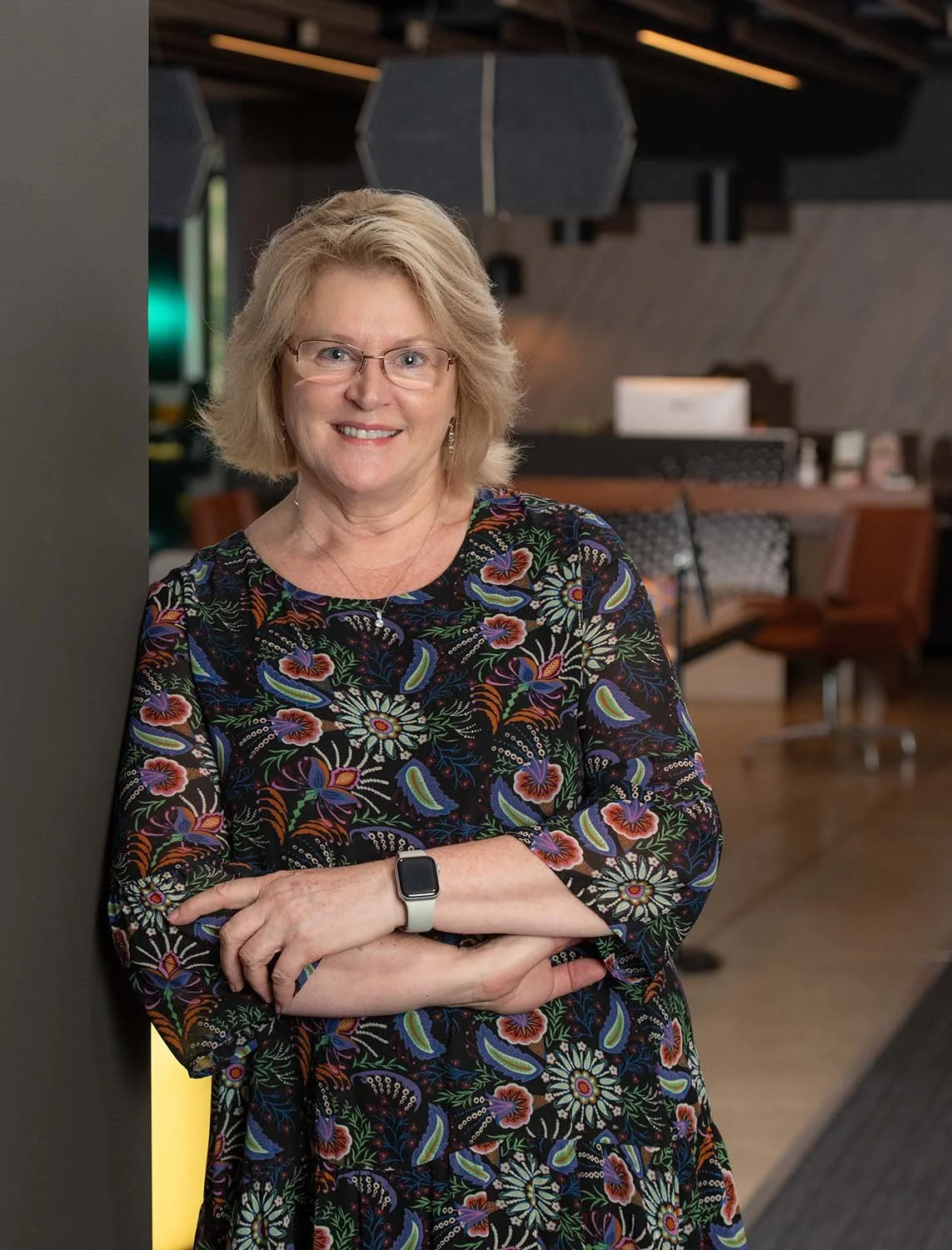 A woman with blonde hair, glasses, and a floral dress leaning against a wall in a modern office or lounge area, smiling at the camera.