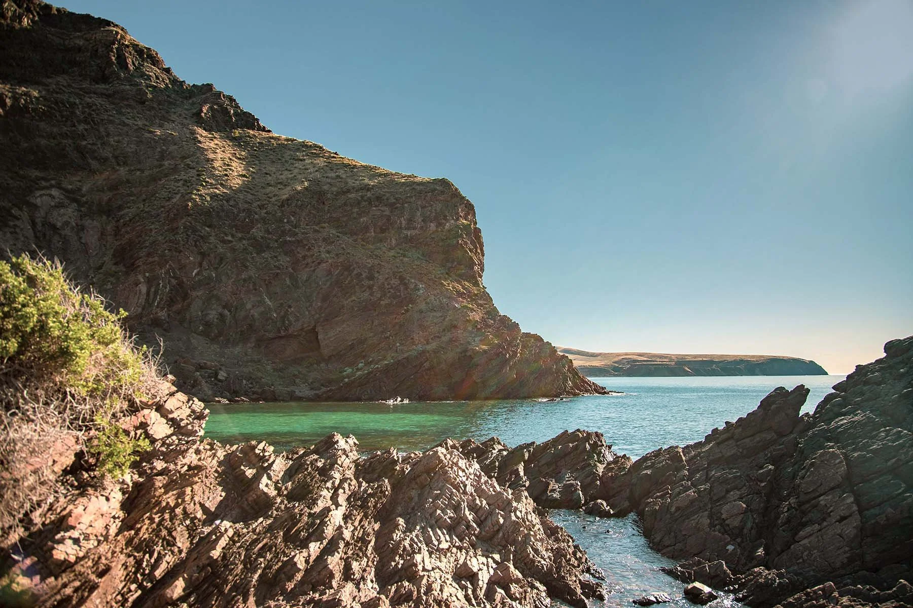 A rocky coastline with cliffs, green water, and clear blue sky.