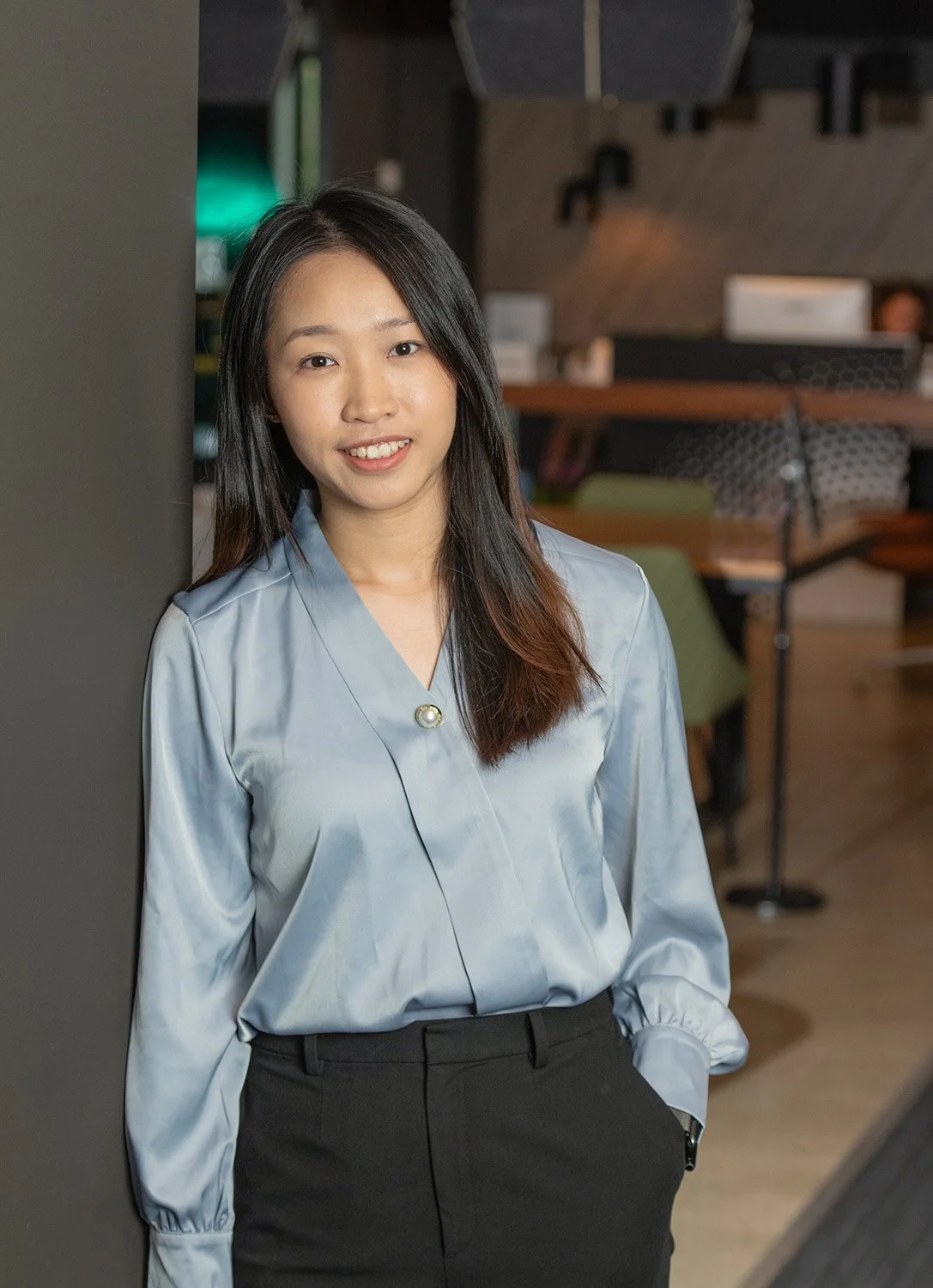 A young woman with long black hair, wearing a light blue satin blouse and black pants, smiling and standing in a modern office or cafe.