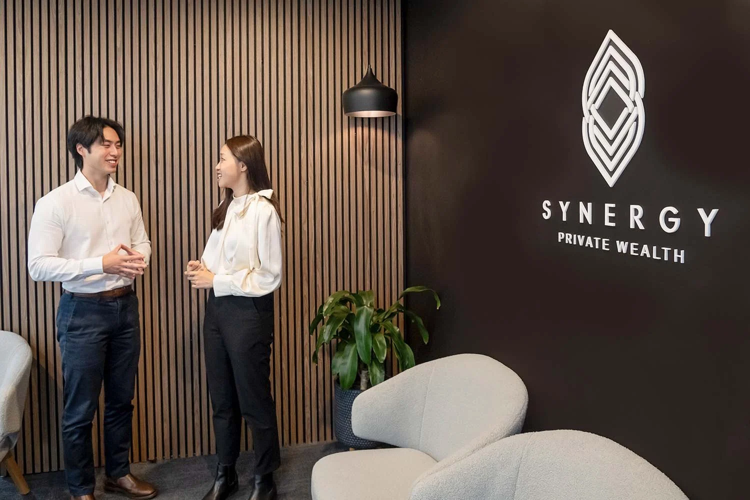 Marven Yap and Krystal Zhong talking and smiling in Synergy Private Wealth Adelaide office lobby. There is a wall with wooden slats, a black wall with a logo and the words 'Synergy Private Wealth,' a potted plant, and modern gray chairs.