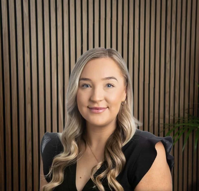 Portrait of a young woman with long wavy blonde hair, wearing a black top, smiling, against a wooden slat background with a plant on the right
